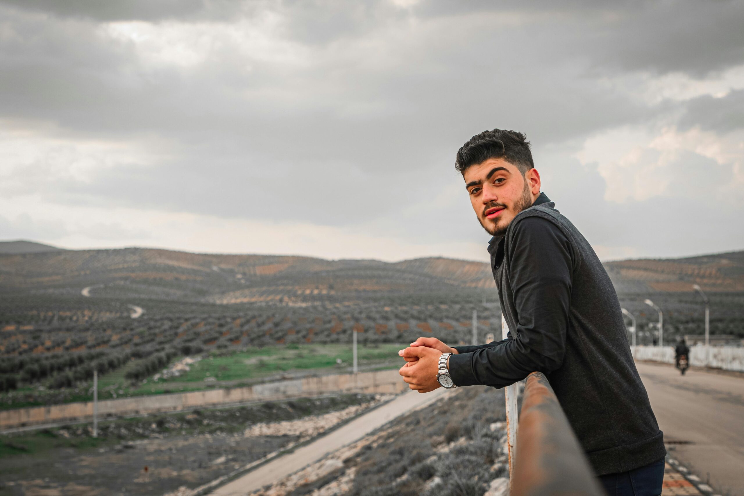 Home Young man in black sweater leaning on a fence, overlooking a vast open landscape. Cloudy sky in the background.
