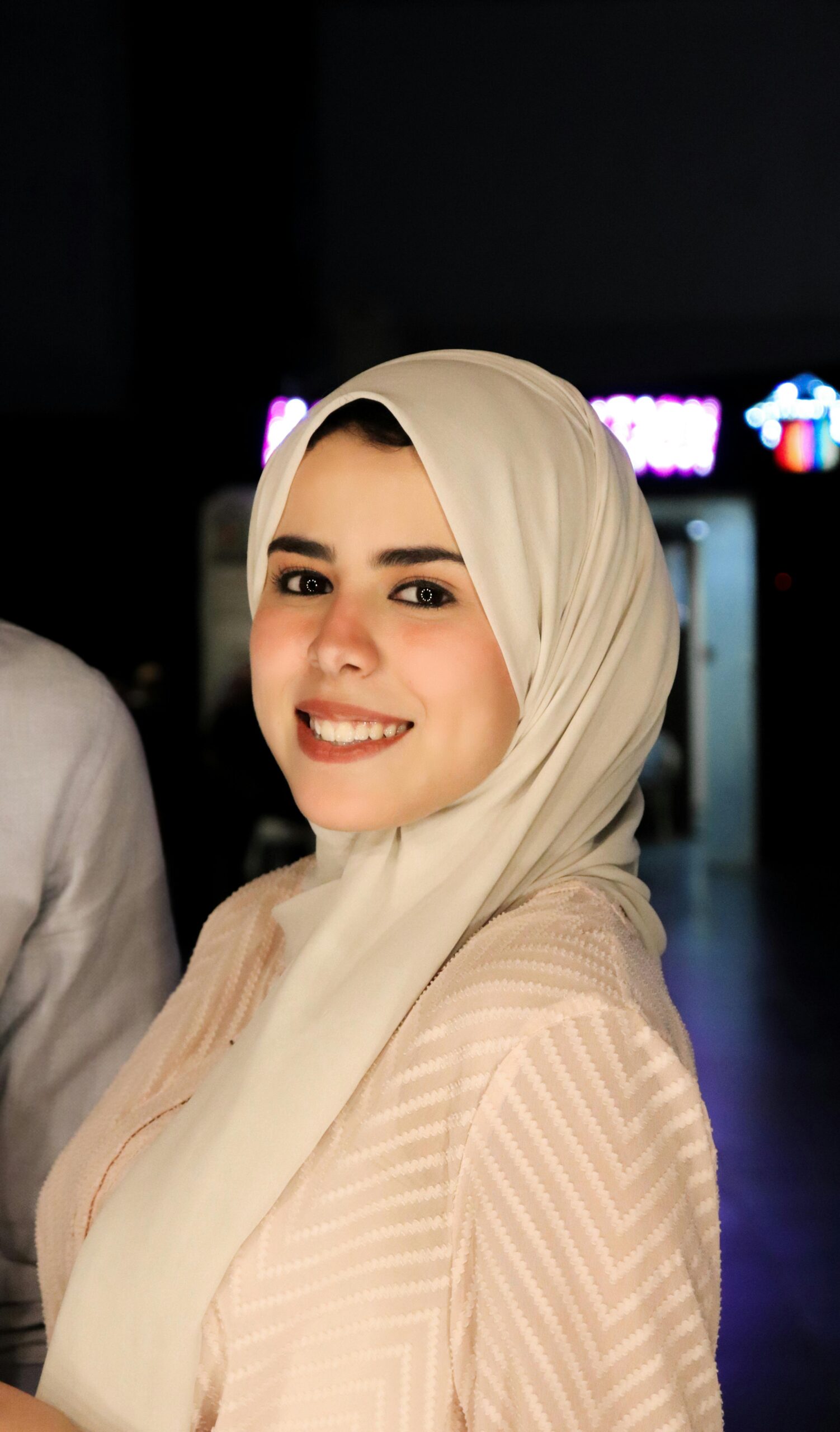 Home A young woman in a hijab smiles warmly at the camera, captured indoors.